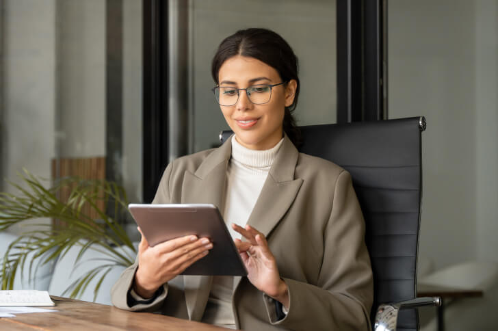 Lady wearing glasses sat in an office chair using and iPad while smiling